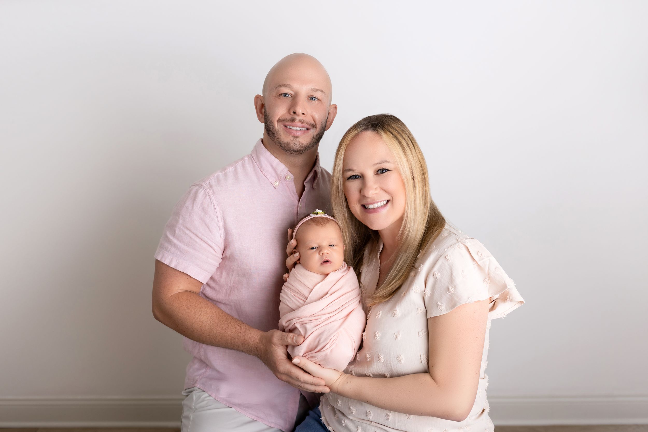 A family in light pink and white clothing poses together against a neutral wall backdrop for a studio portrait.
