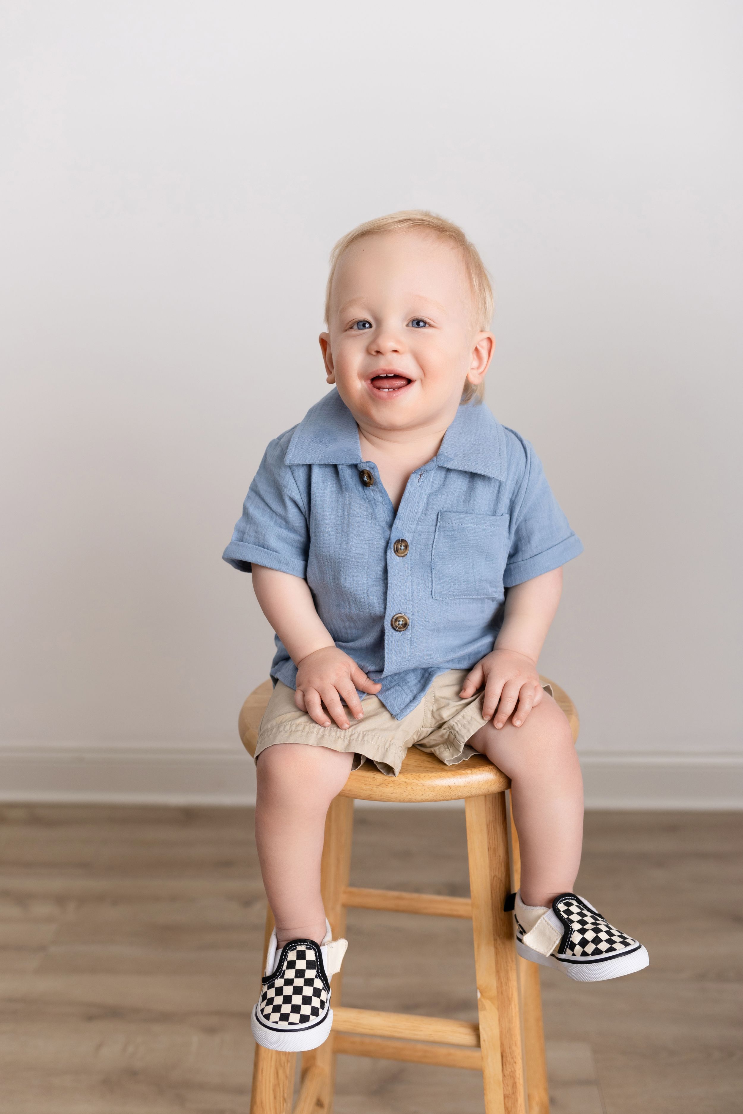Baby wearing blue collared shirt and checkered slip-on shoes sits playfully on wooden stool showing different expressions.