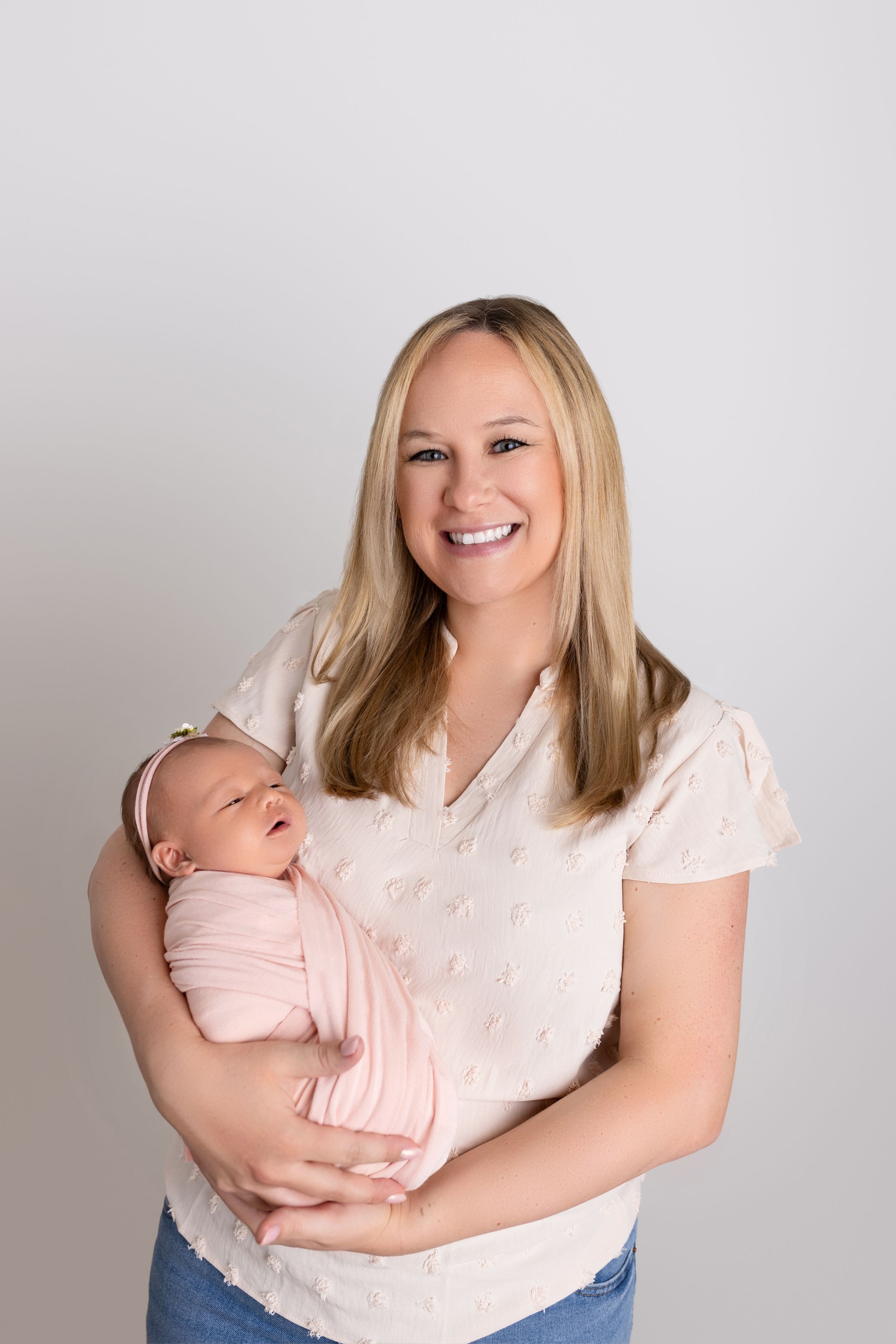 A woman in a light pink blouse holds a sleeping newborn baby against a neutral studio background.
