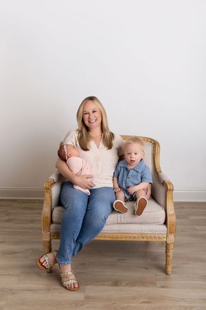 A parent and baby in matching light colors sitting together on a wooden rattan settee in a minimalist setting.