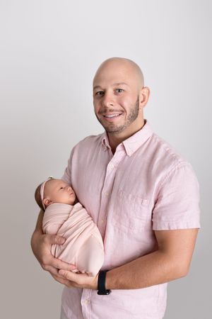A parent in pink shirt holding and kissing a newborn baby against a neutral background.