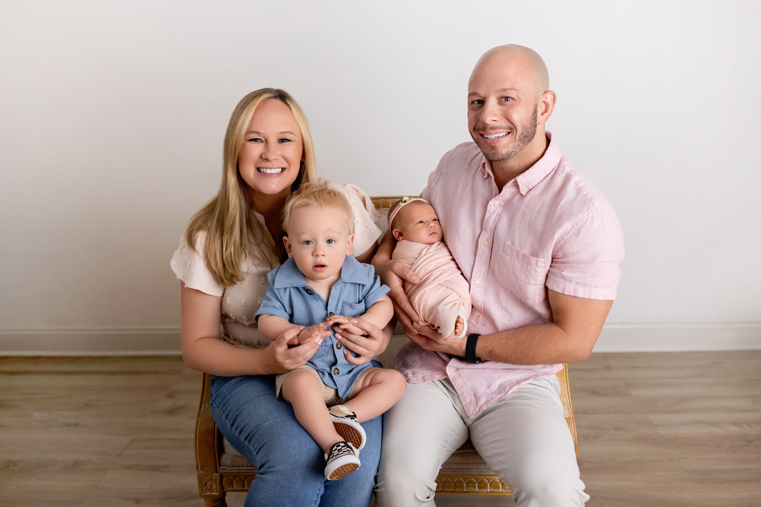 Family sitting together in casual attire on hardwood floor against light colored wall for professional portrait.
