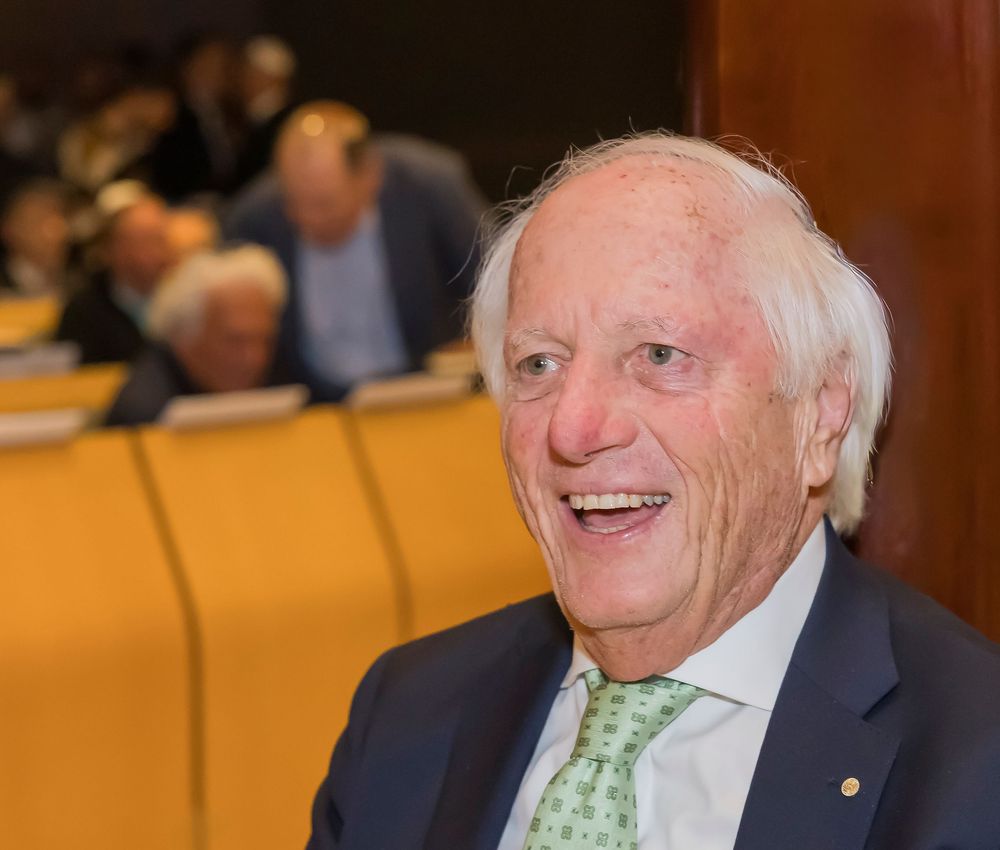 An elderly man wearing a navy suit and green tie smiles warmly while seated in an auditorium setting.