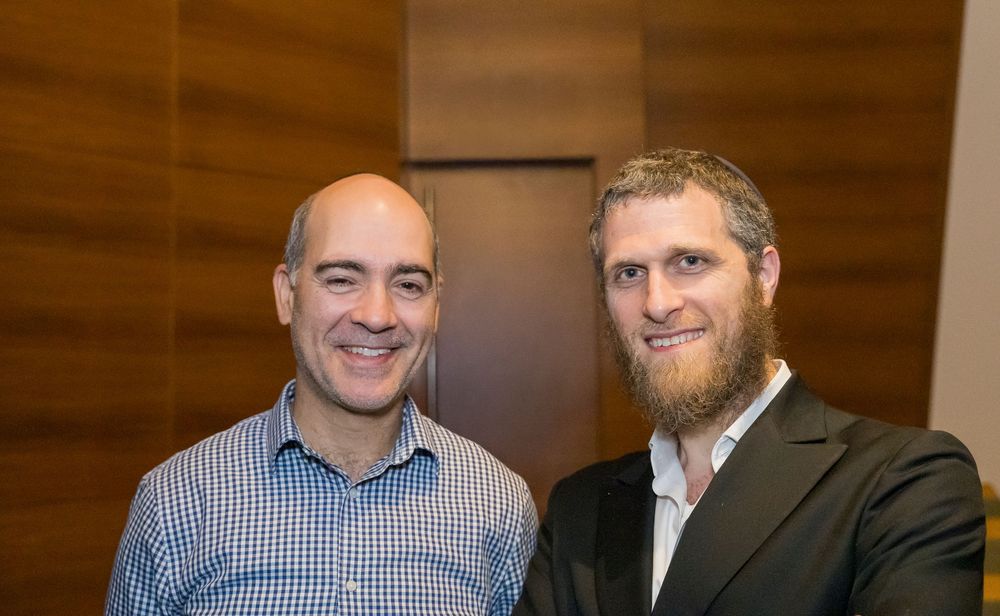 Two people posing together at an event, one wearing a checkered shirt and one in a dark blazer with a beard.