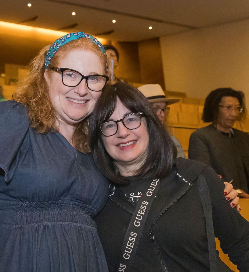Two people wearing glasses smile together at an event in a warmly lit indoor venue.
