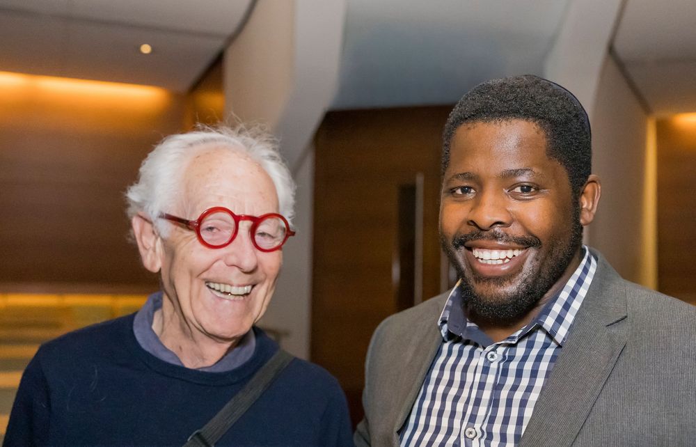 Two men smile together at an indoor event, one wearing red glasses and one in a gray suit jacket.