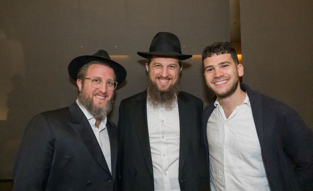 Three men pose together at a Jewish event, two wearing traditional black hats.