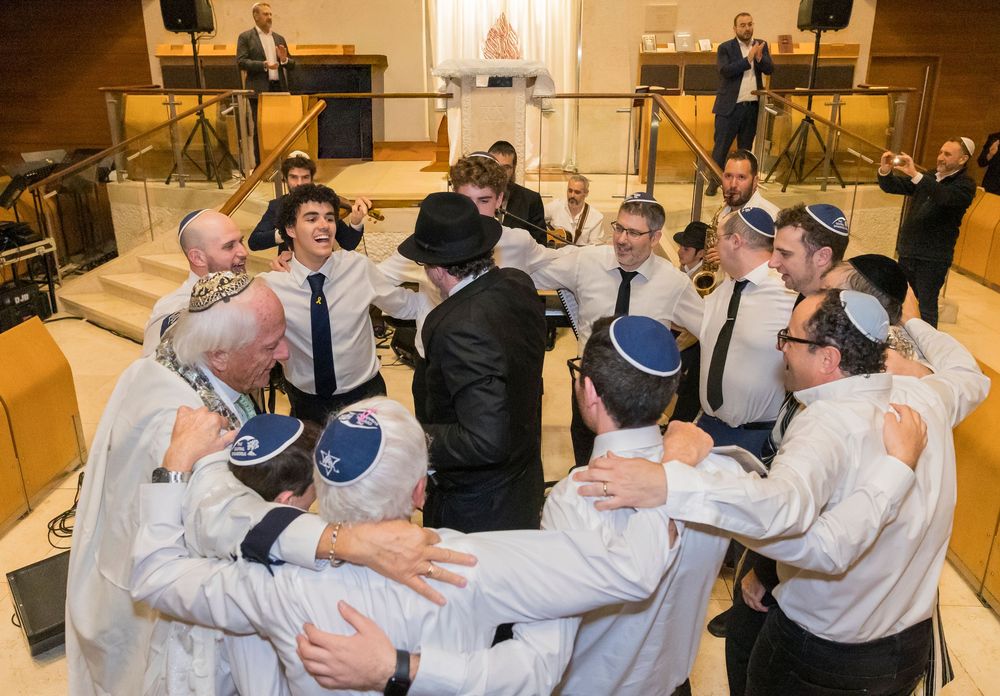 A group of men in kippahs and formal attire huddle together in celebration at a synagogue event.