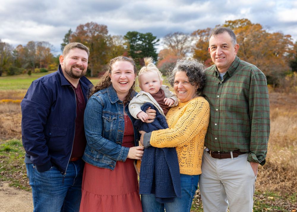 3 generations of a family pose outdoors in autumn with fall foliage and natural scenery in the background in Concord, MA.