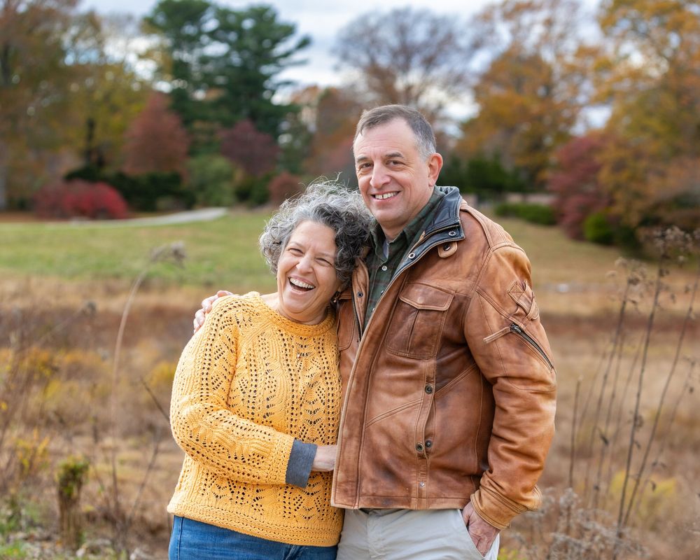 A couple wearing a yellow knit sweater and brown leather jacket embrace outdoors with fall foliage during multi-generational photos Concord.
