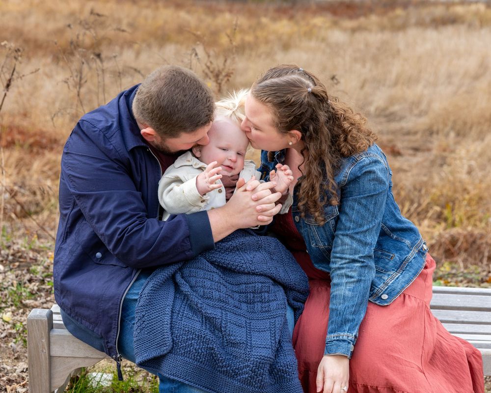 A mom and dad tenderly embrace their baby at outdoor photos against a natural autumn landscape background during multi-generational photos