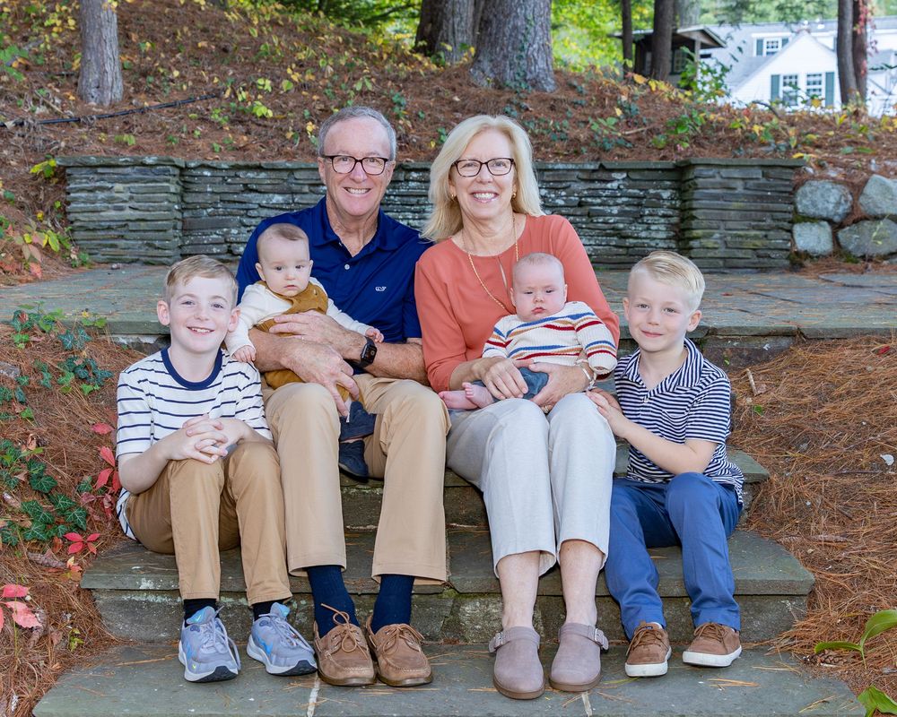 Granparents and grandchildren pose together outdoors on stone steps surrounded by trees and autumn leaves in a casual portrait setting.