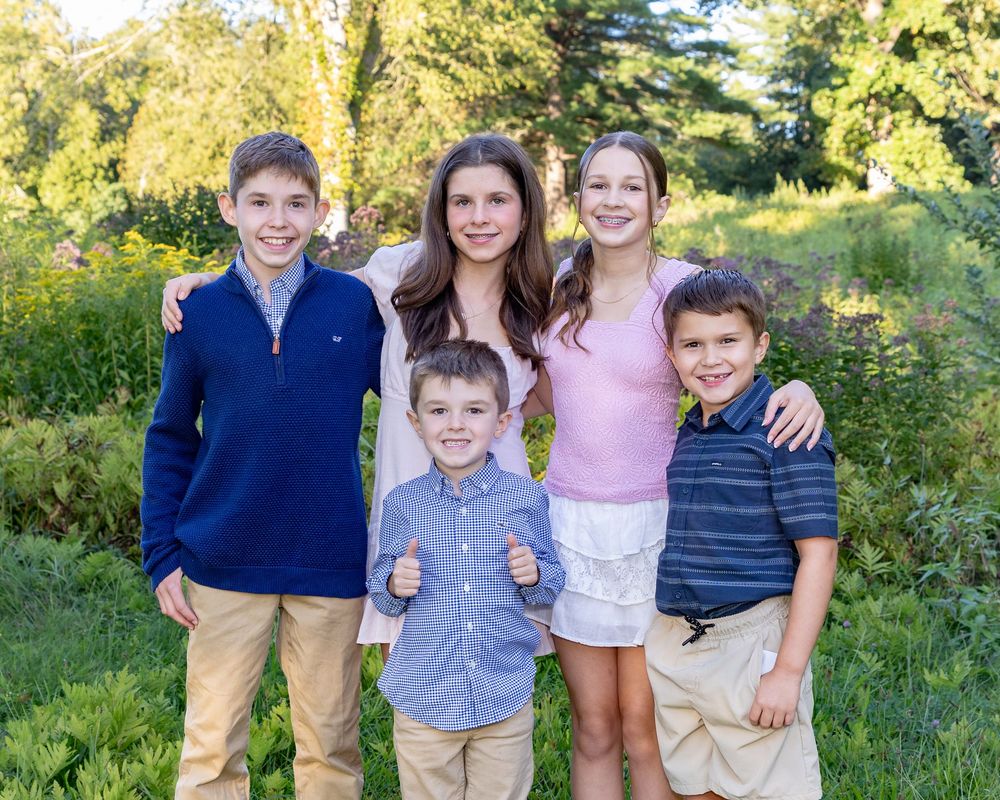 Cousins pose together outdoors in casual clothing against a sunlit natural backdrop with green foliage at North Bridge in Concord, MA.