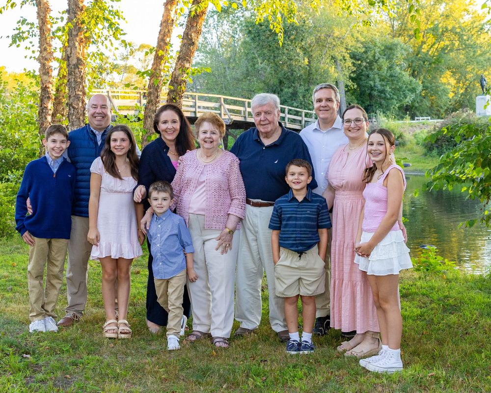 Multi-generation family in coordinated pink and blue outfits poses together outdoors in a natural setting during golden hour in Concord, MA.