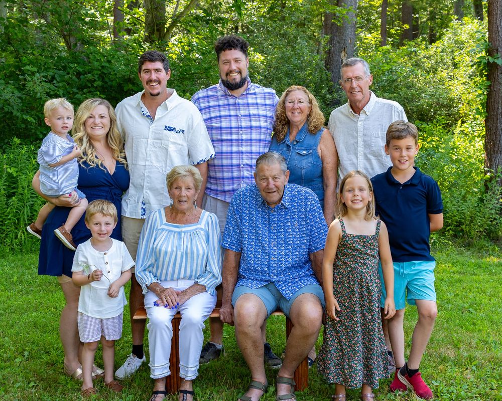 Four generations of a family poses together outdoors in a lush green garden setting during a sunny summer day in Acton, MA.