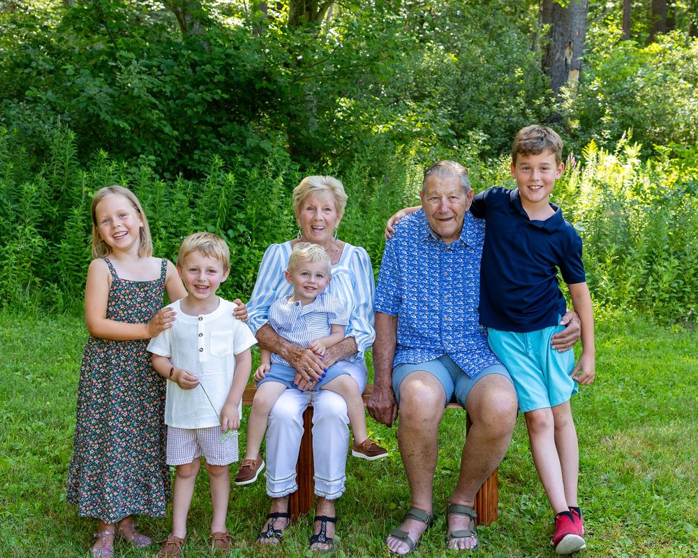Great grandparents and children pose together on a wooden bench surrounded by lush green foliage in summer during extended family photos.