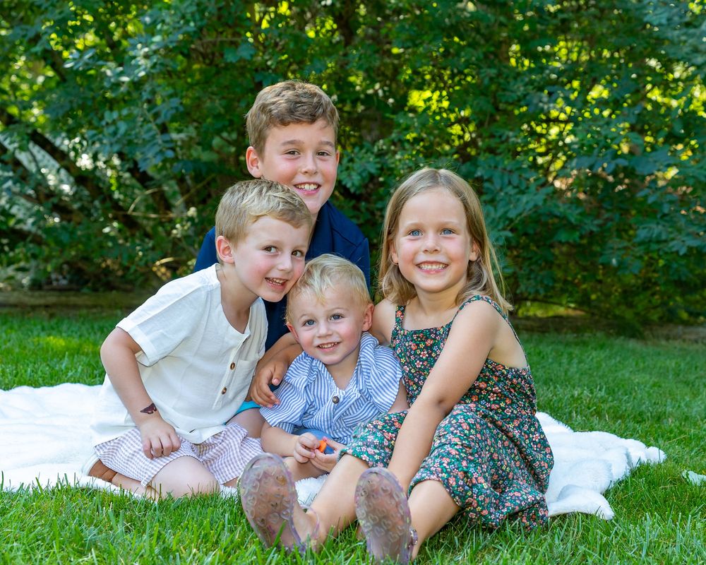 Cousins sitting together on a white blanket in a green grassy area with trees in the background during extended family photo session.