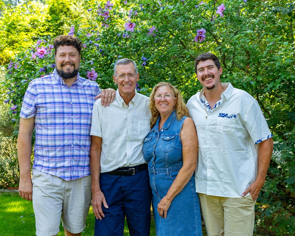 Parents & adult children pose outdoors in casual attire with lush green foliage and purple flowers in the background in Acton, MA.