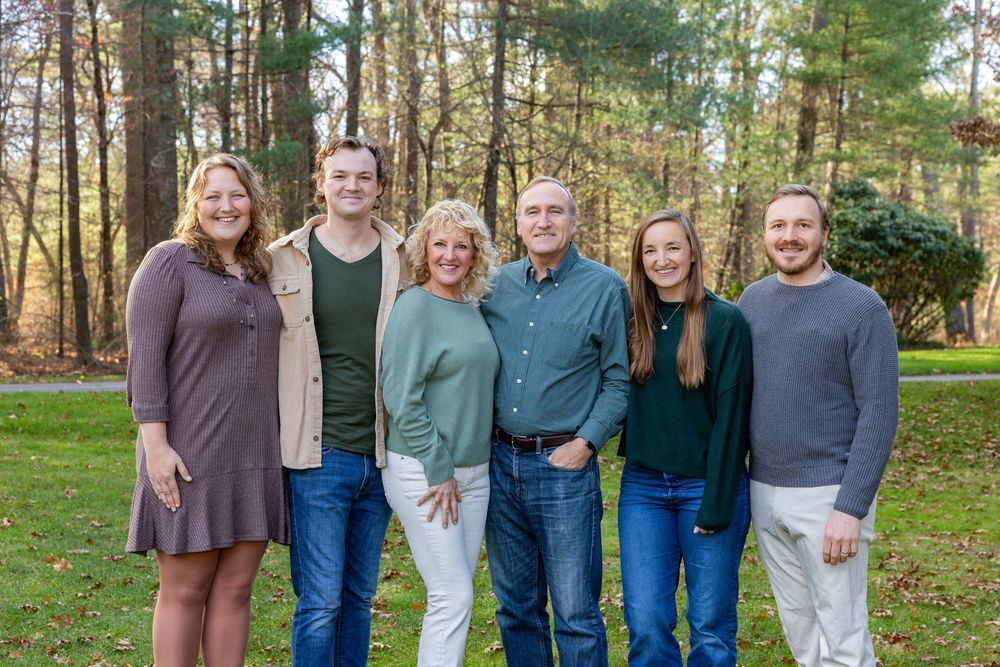 Parents and their adult children pose together in a wooded setting with pine trees in the background during multi-generational family photo.