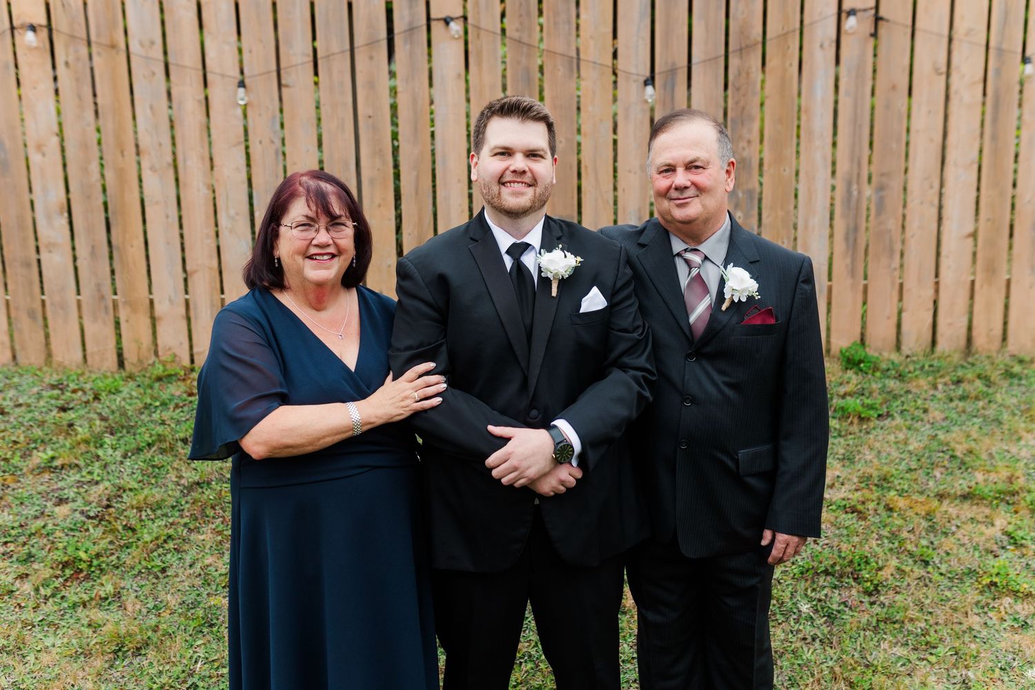 Wedding party members in formal attire pose against wooden fence backdrop during outdoor celebration.