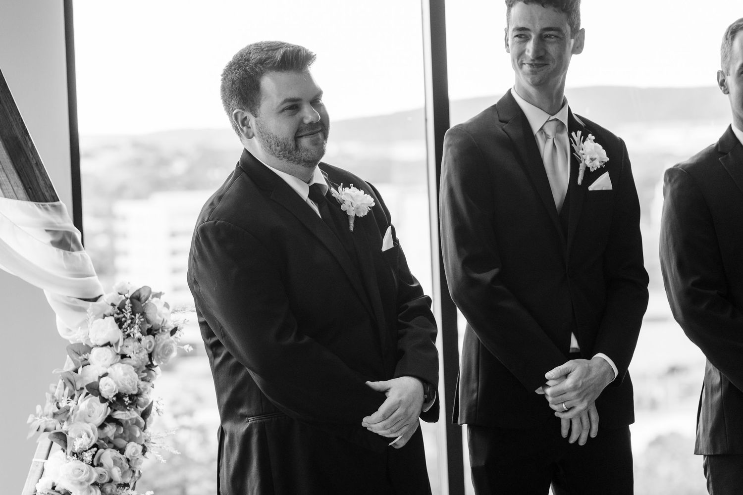 Wedding party members in dark suits stand together laughing outdoors.