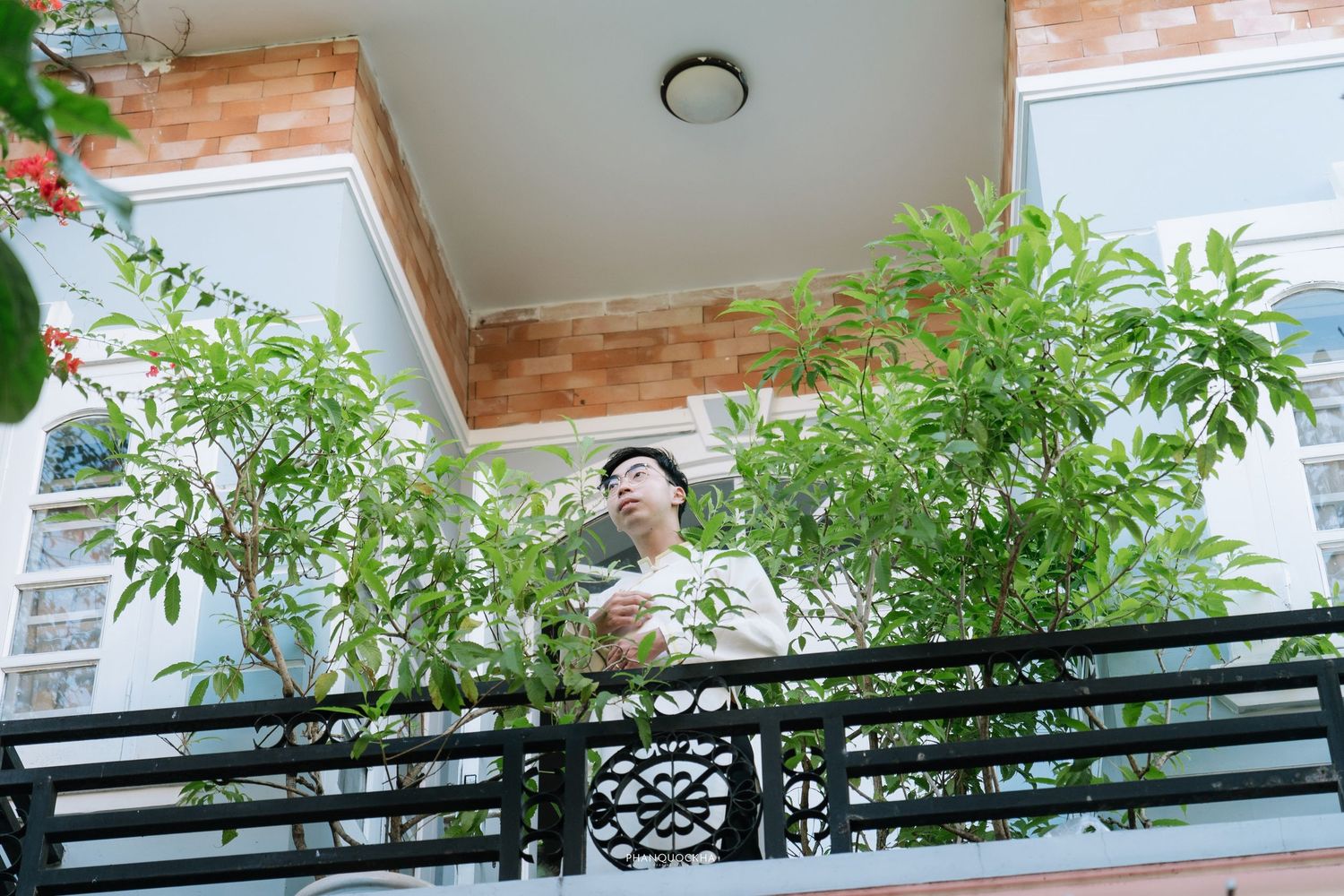 Groom in white ao dai stands on flower-adorned balcony looking down at wedding venue below.