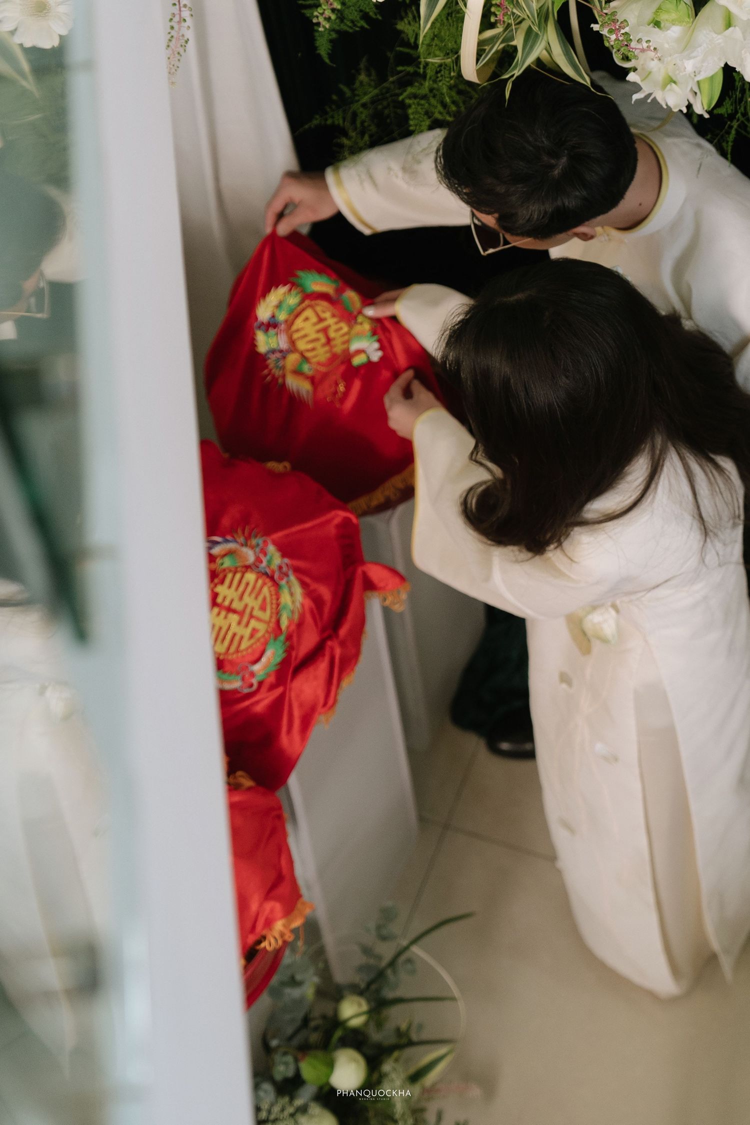 Couple in white ao dai arranging red embroidered wedding gift boxes during Vietnamese engagement ceremony.