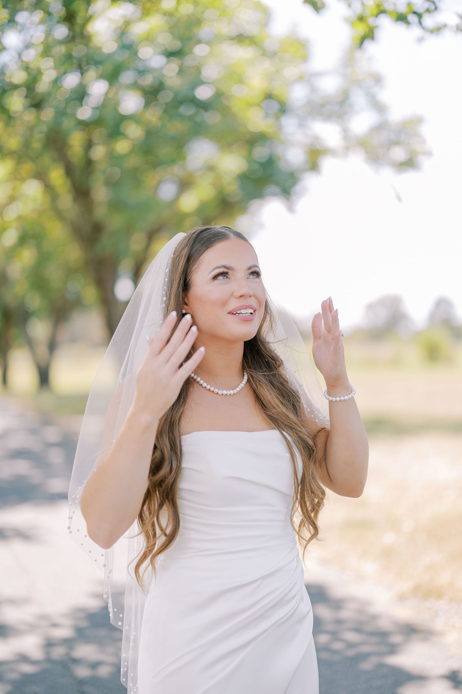 A bride in a white wedding dress smiles while adjusting her veil outdoors on a sunny day.