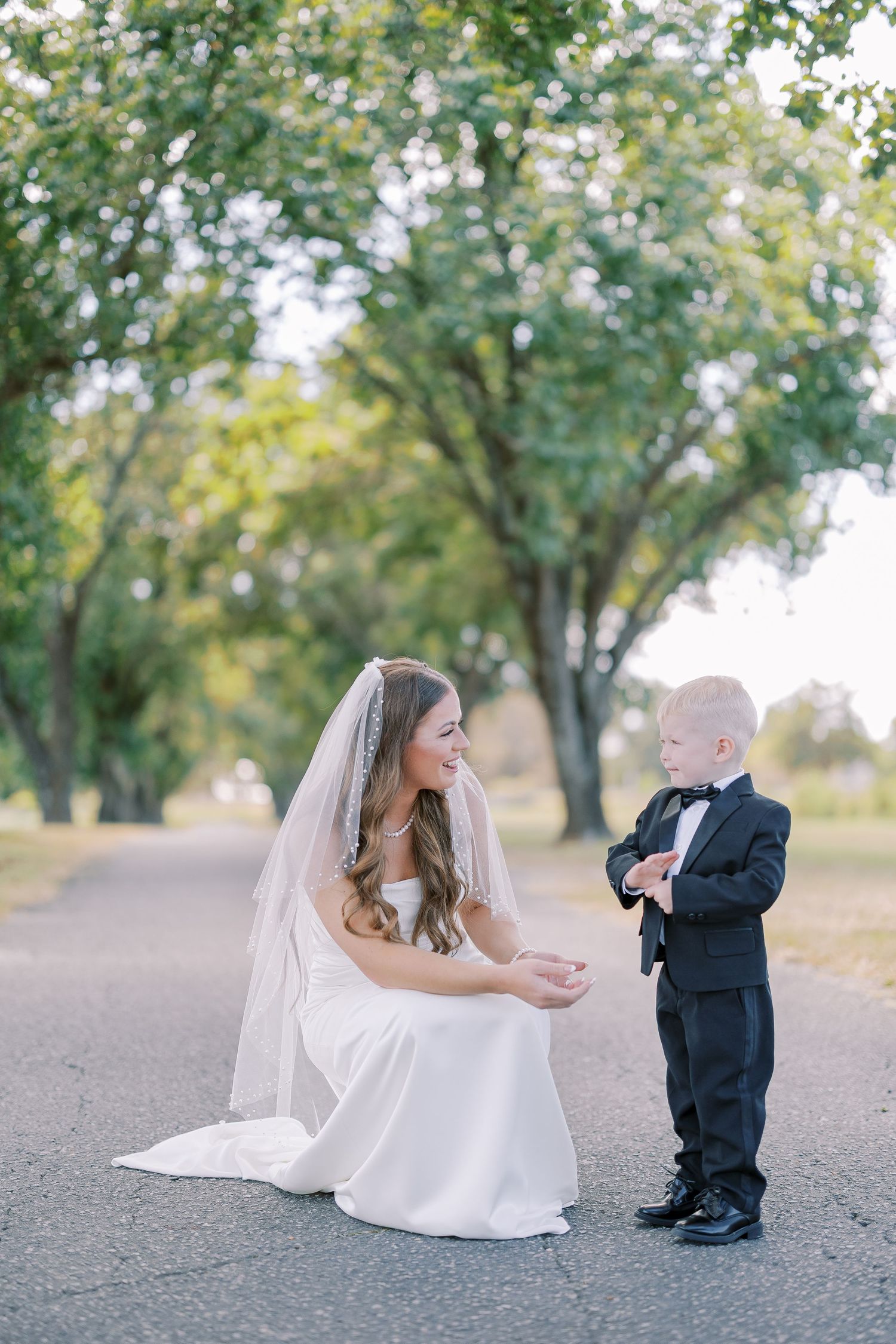 A bride in a white dress crouches down to interact with a young child dressed in formal attire at an outdoor photo session.