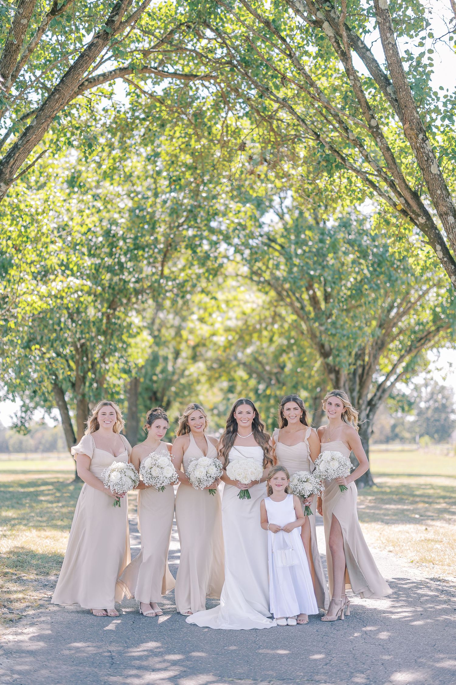 Multiple bridesmaids in blush-colored dresses pose with bride in white gown under shady trees on sunny day.