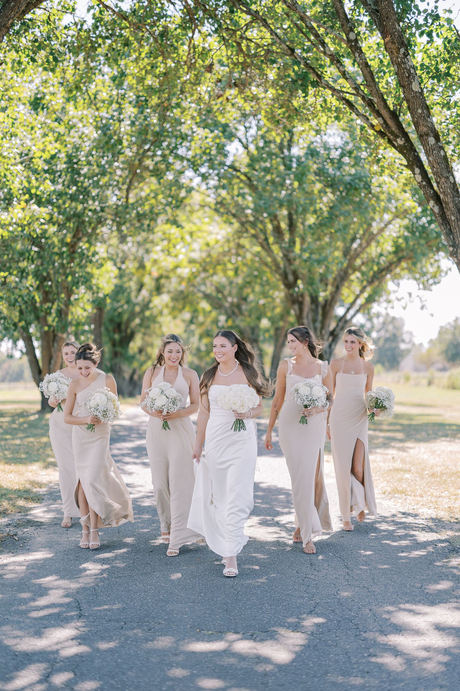 Bridal party in blush dresses walking and laughing together on a sunlit tree-lined path for outdoor wedding photos.