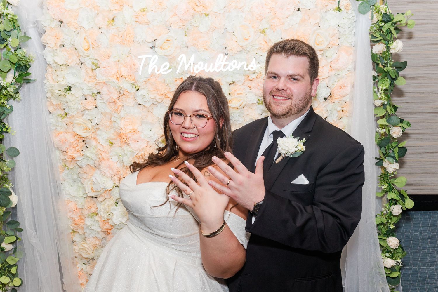 A celebratory moment at a wedding with a floral wall backdrop in soft peach and white tones.