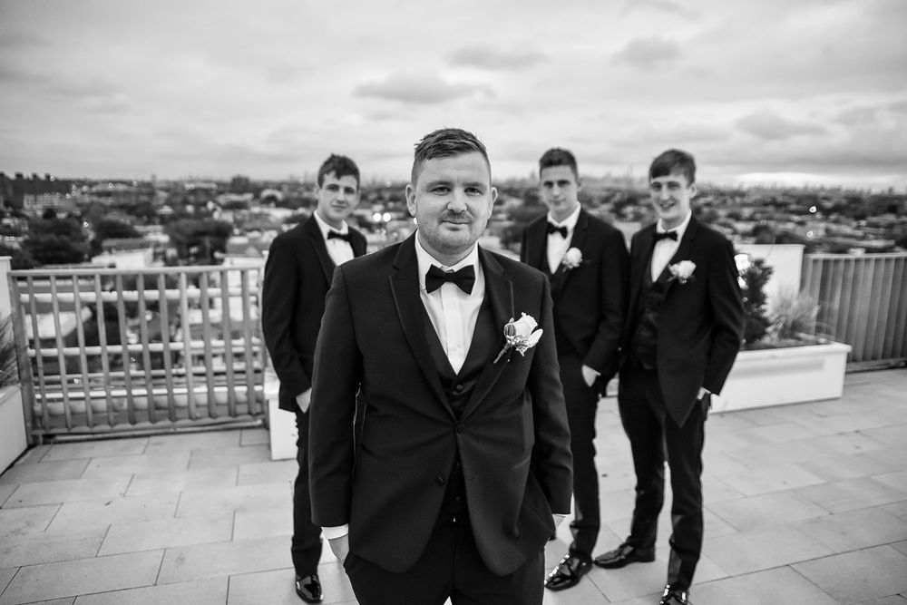 A wedding party in black tuxedos poses for photos on a rooftop with city views in the background a