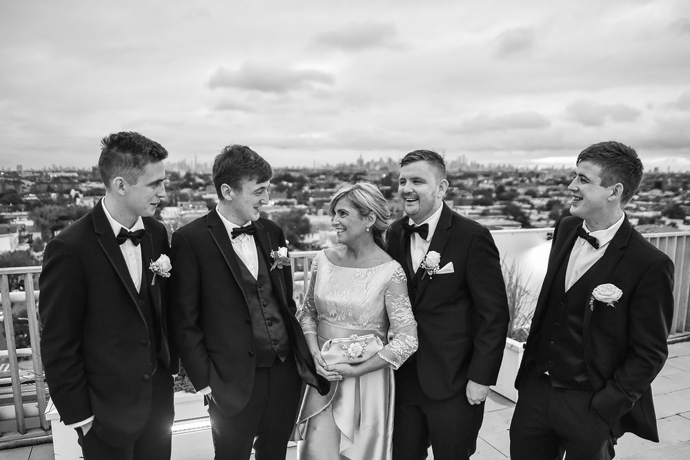 A wedding party in formal attire stands together on a rooftop overlooking a cityscape on an overcast day.