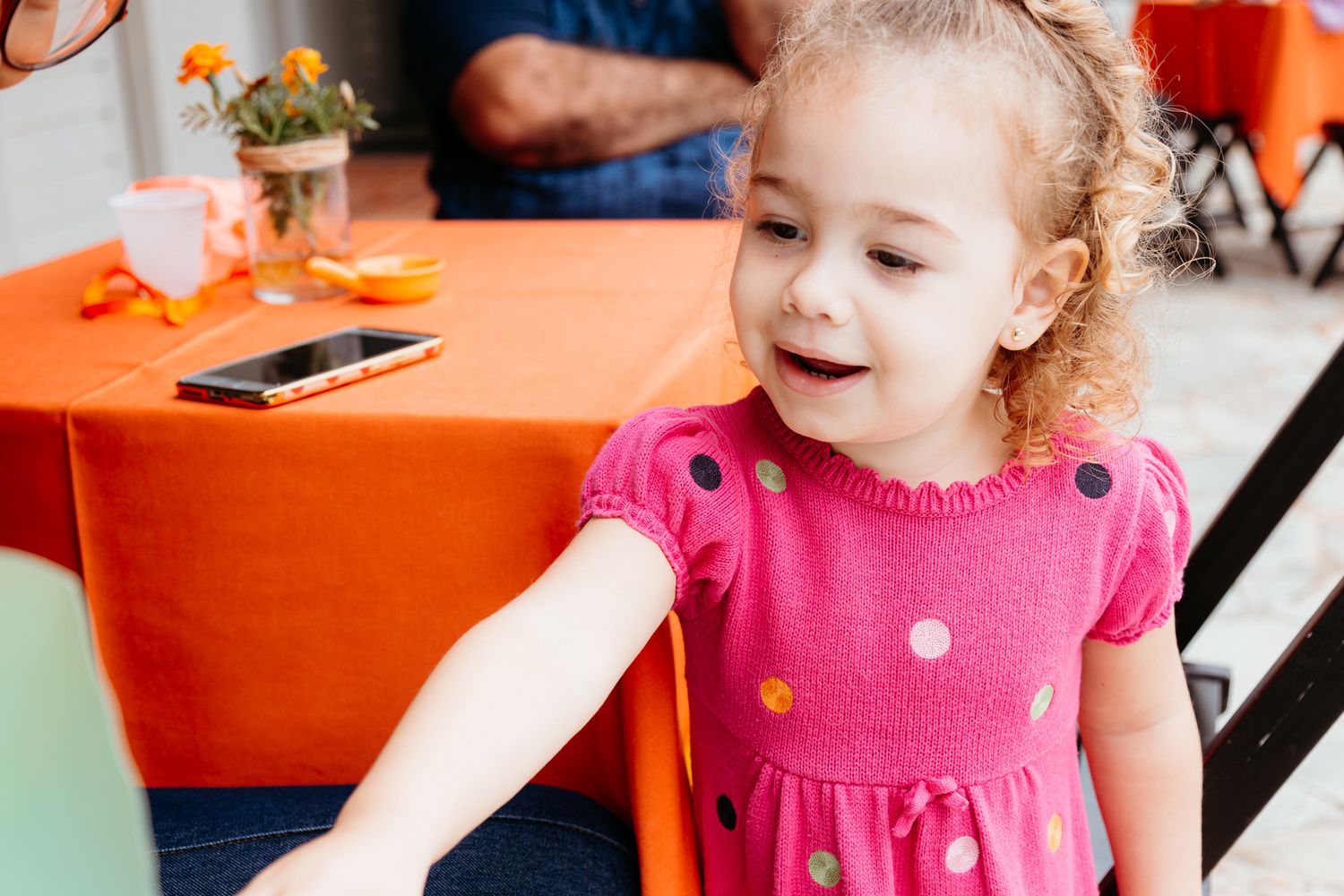 A child in a bright pink polka dot dress sits at a table with an orange tablecloth.
