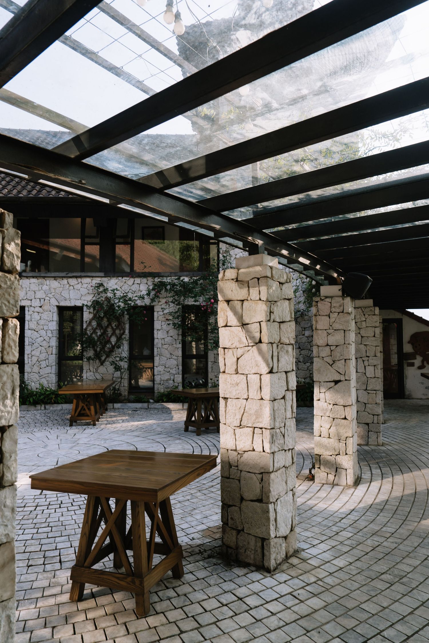 Stone pillar courtyard with wooden tables under transparent pergola roof.