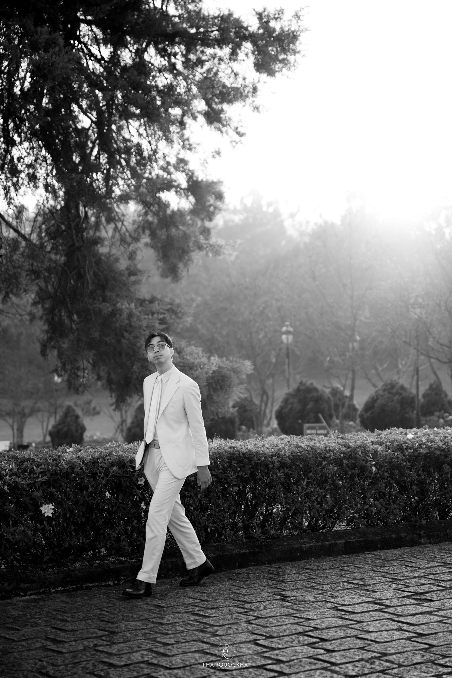 Man in white suit walking along a hedge-lined brick path in a misty park, black and white photo.