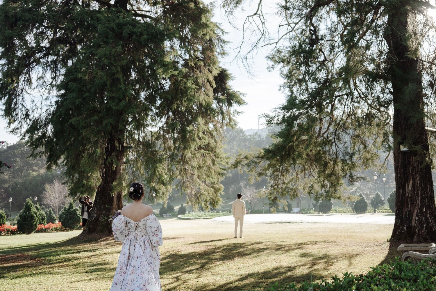 Bride and groom standing apart in a lush green park beneath large trees on their wedding day.