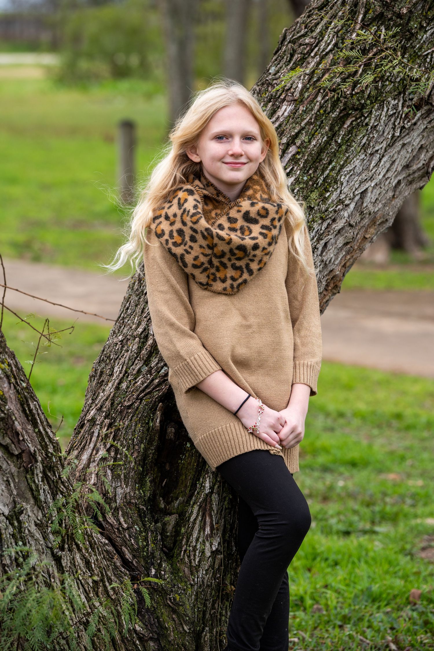 Person in tan sweater and leopard print scarf poses by tree trunk in outdoor nature setting.