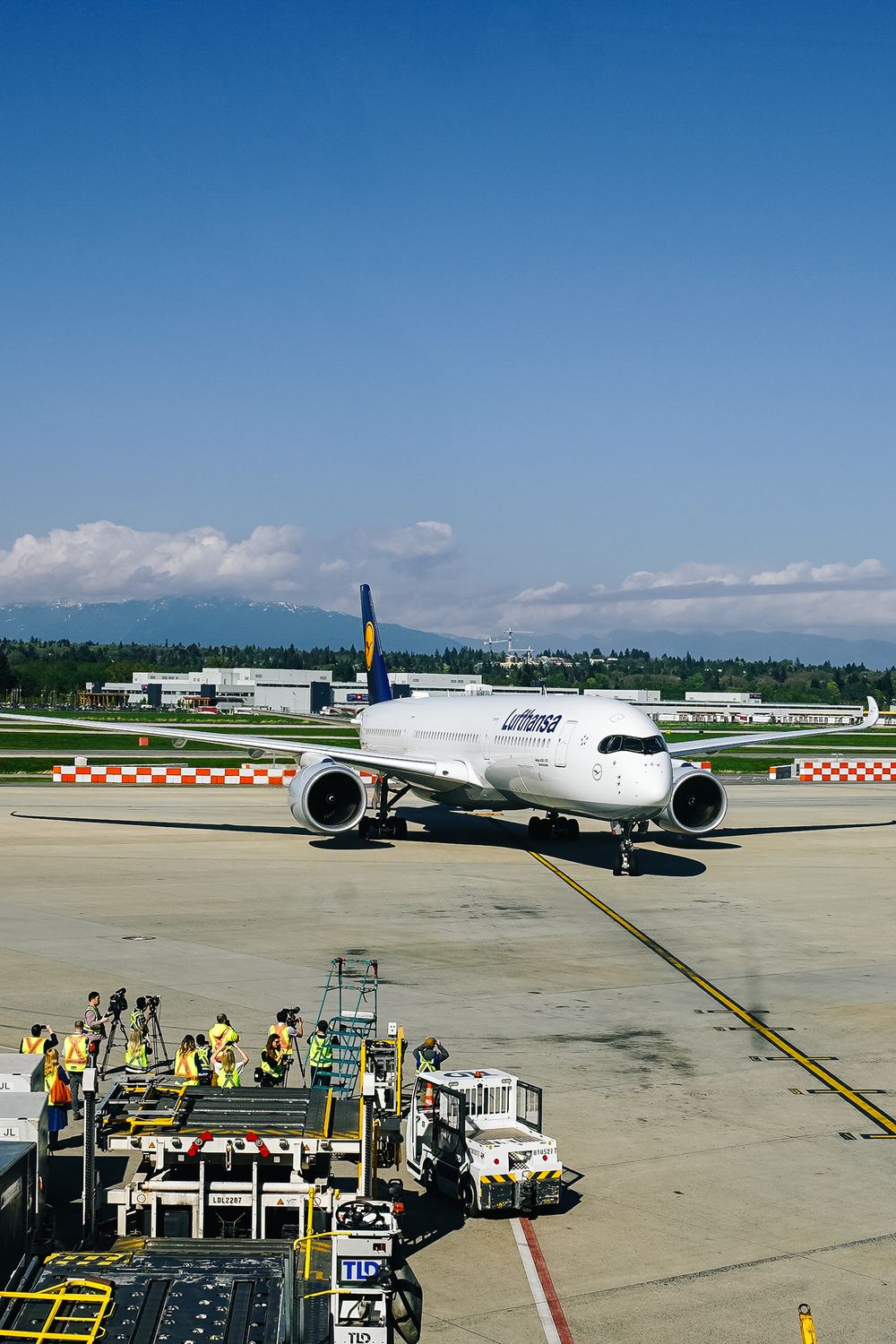 Commercial aircraft parked at airport gate with ground crew performing maintenance and service operations on sunny day.