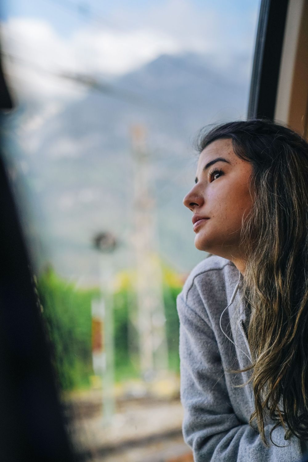 A woman in a gray sweater leans against a window on a cloudy day, gazing thoughtfully into the distance.