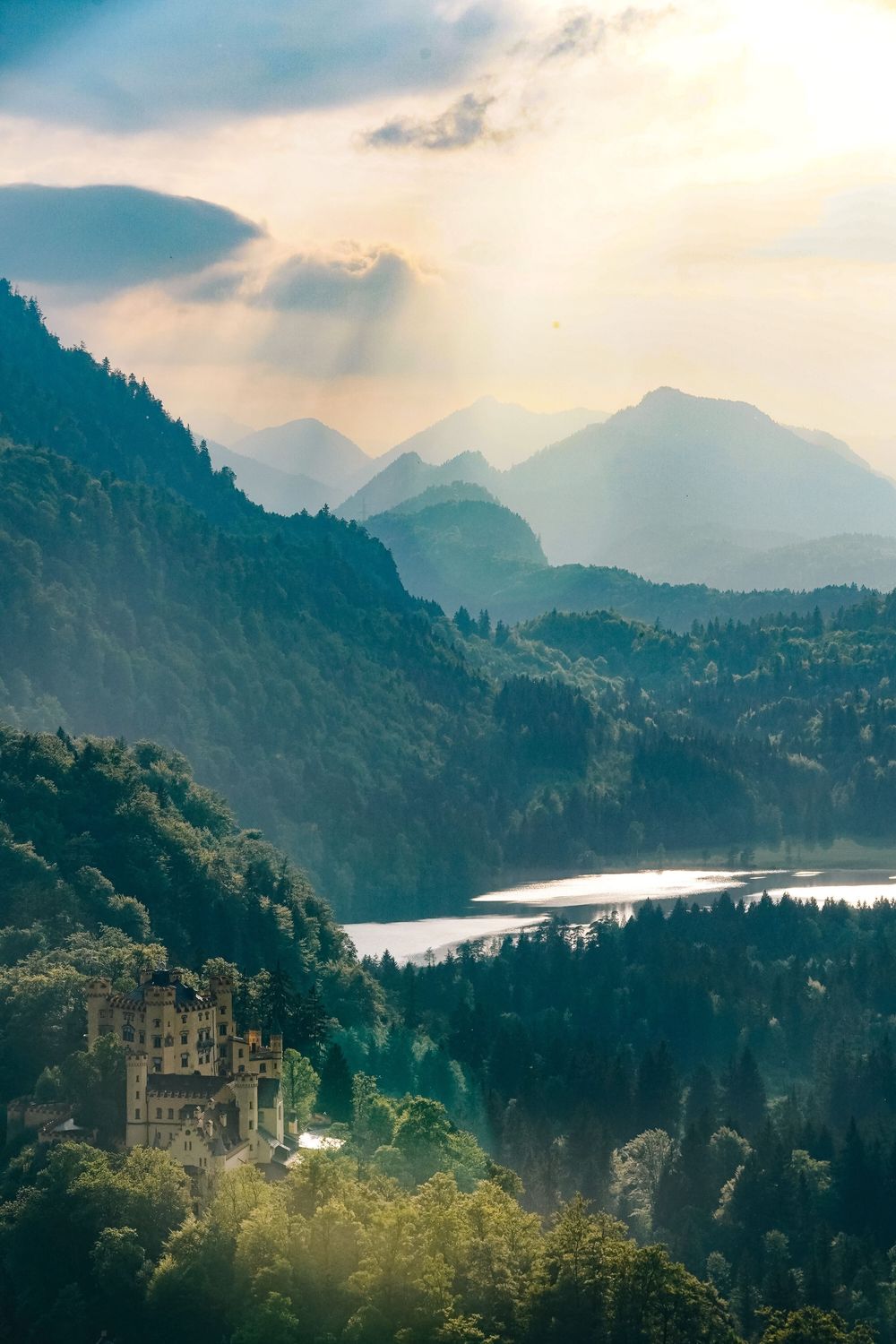 Panoramic view of misty mountain peaks and a serene lake nestled among forested hills during golden hour.