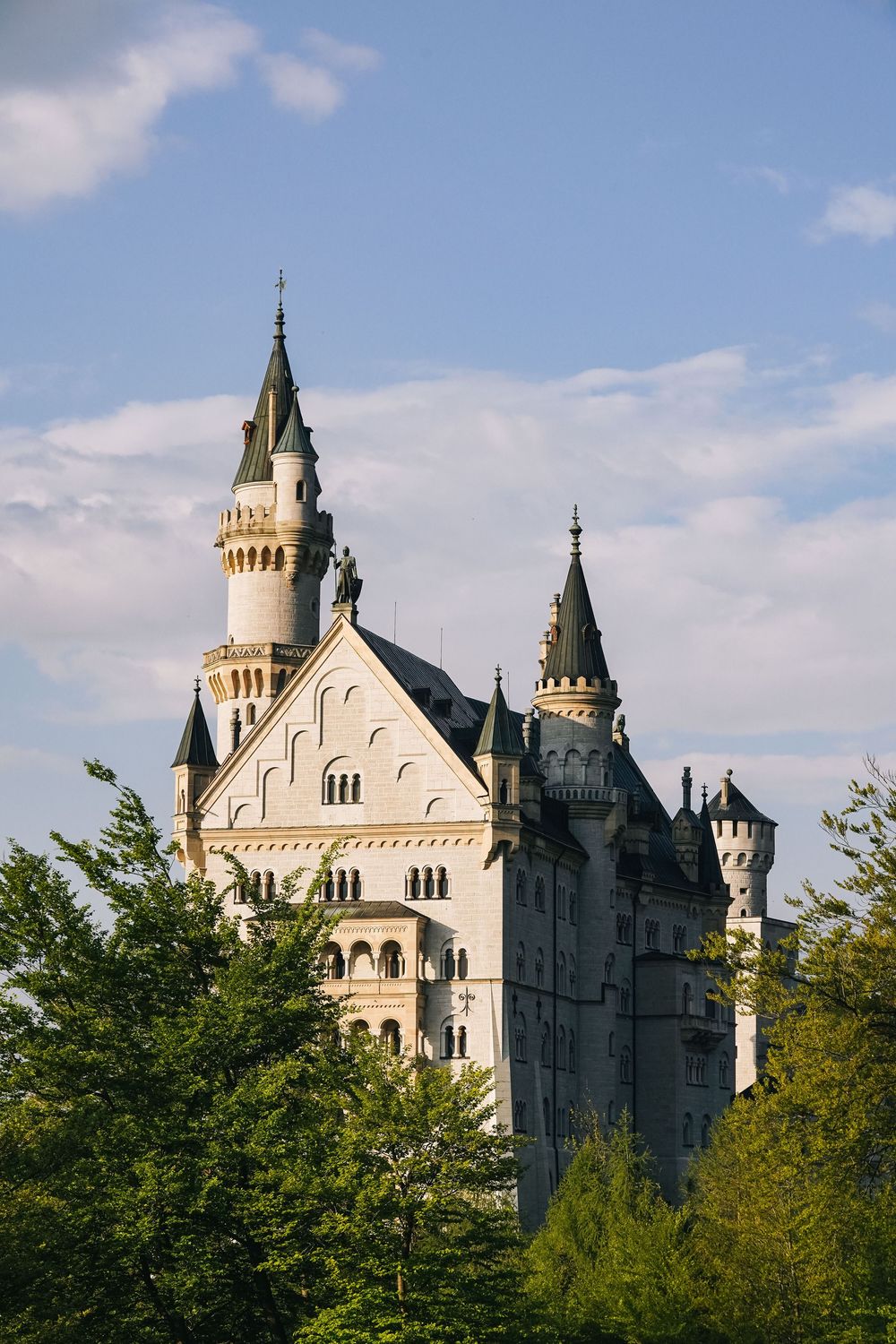 A majestic white stone castle with spires and towers stands against a bright blue sky with trees in the foreground.