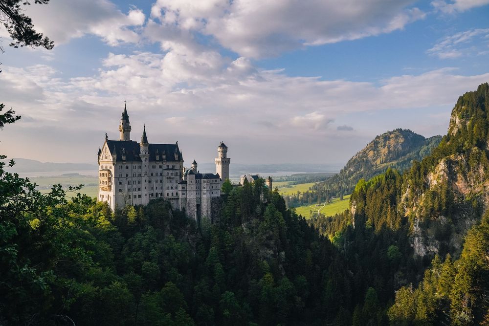 Five panoramic views of Neuschwanstein Castle perched dramatically on a hilltop in the Bavarian Alps under cloudy skies.