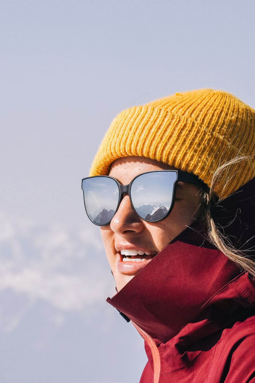 Person in a red winter jacket and yellow beanie hat smiling against a bright sky background while wearing sunglasses.