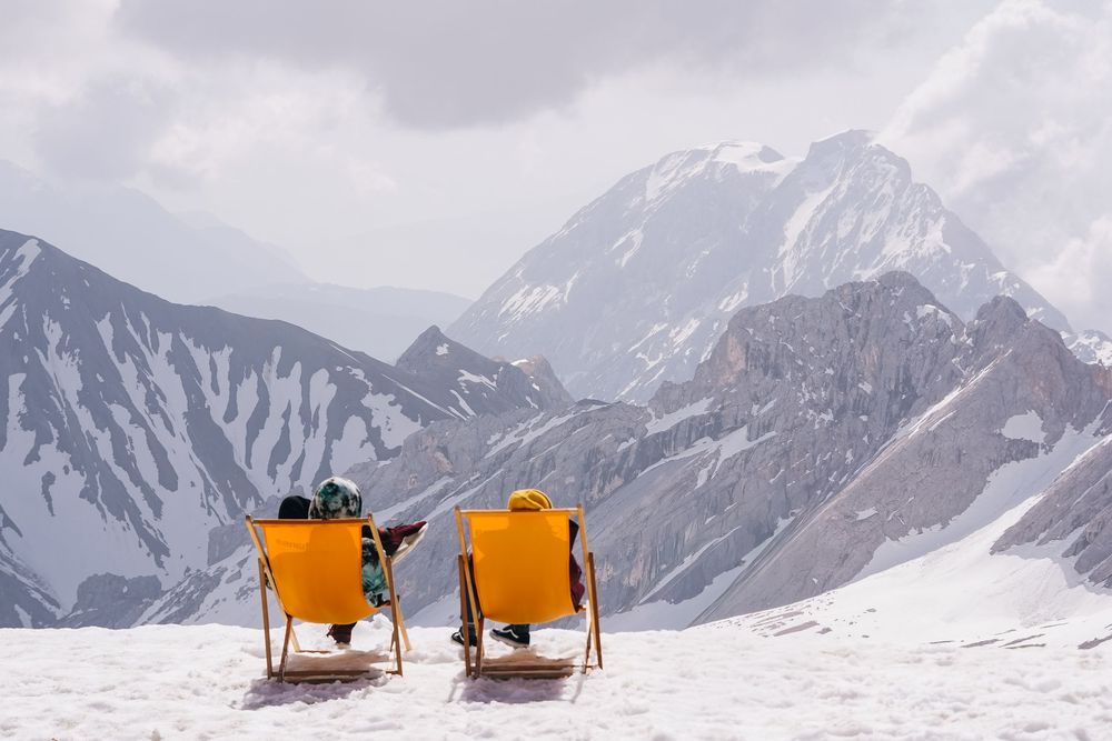 Two orange lounge chairs sit atop a snow-covered mountain peak with dramatic mountain ranges in the background.