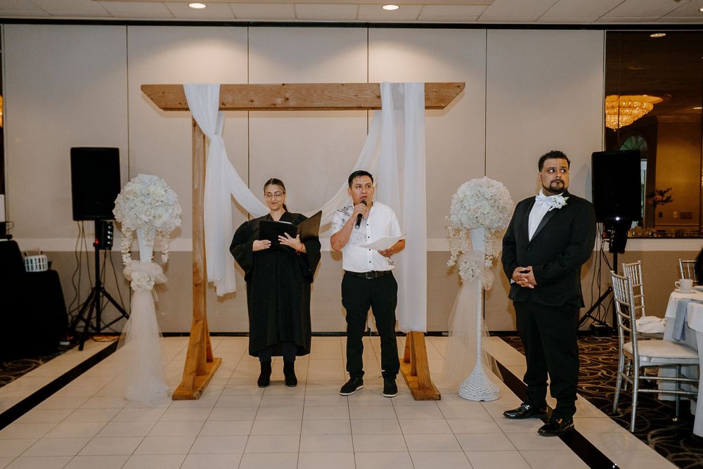 Wedding venue staff members stand by a wooden arch with white floral decorations and audio equipment in the background.