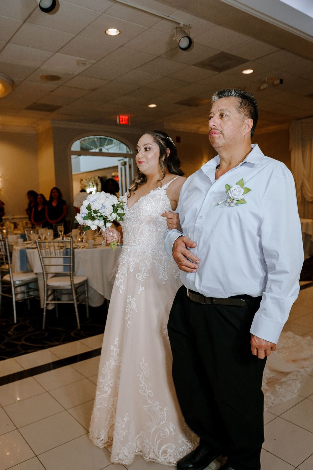 A bride wearing a sparkly white gown stands arm-in-arm with her companion in a wedding reception venue.