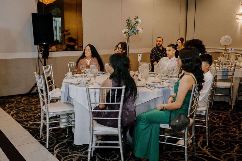 People seated at elegant white-clothed dining tables during an event in a banquet hall with a carpeted pattern floor.