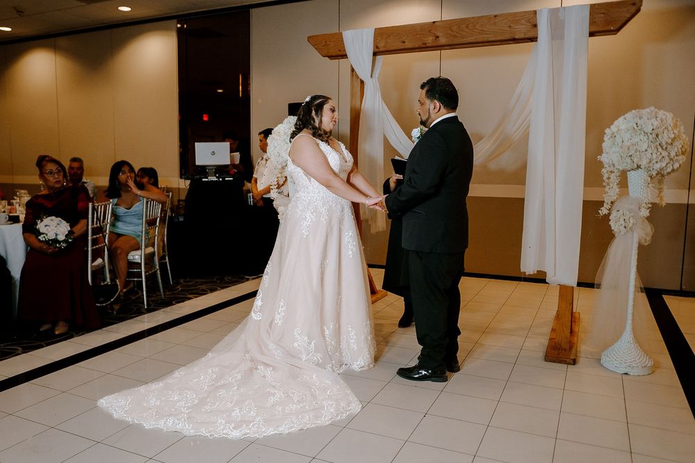 A bride in a white lace wedding dress stands in front of a full-length mirror in a bridal preparation room.
